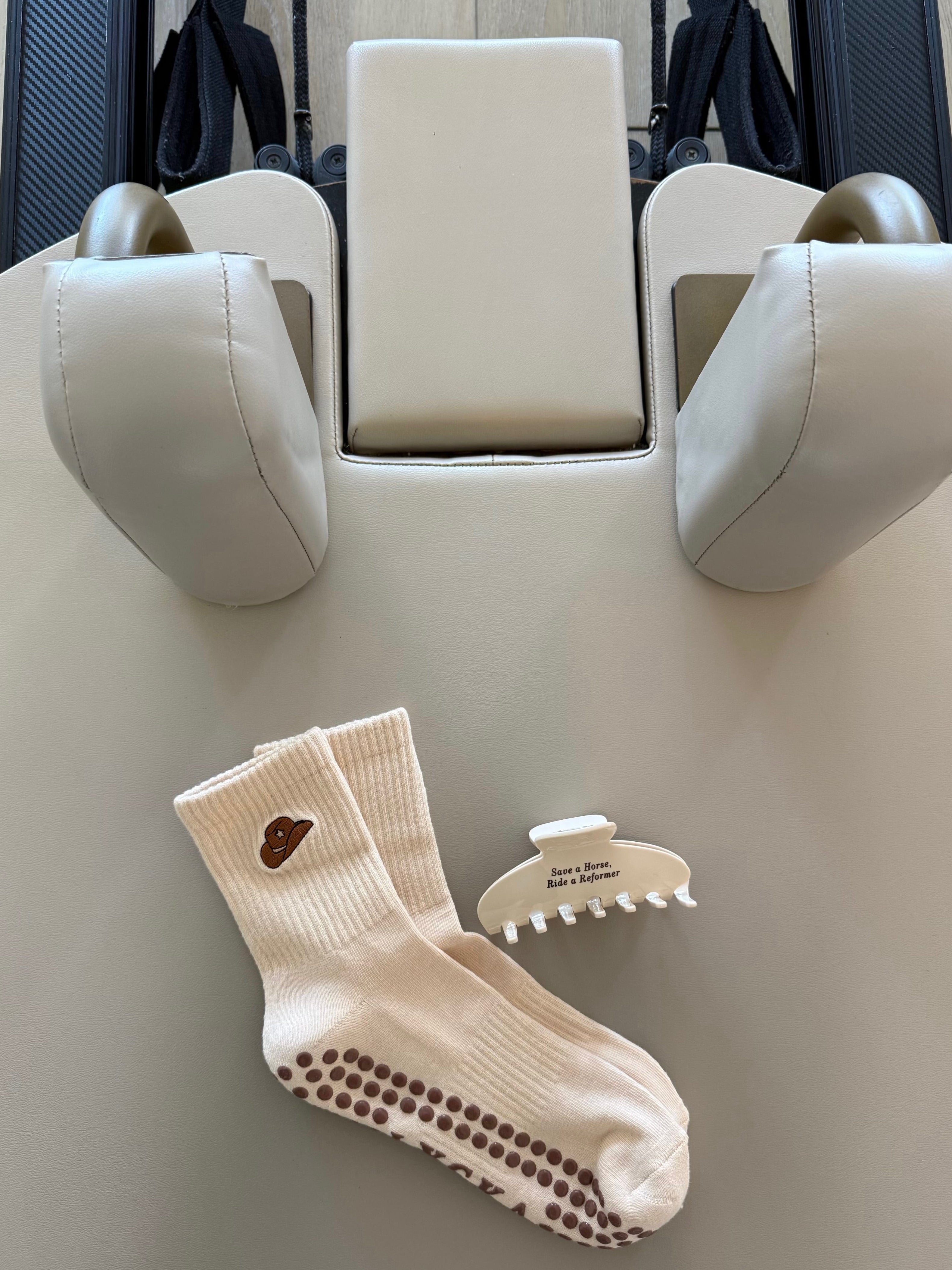 Beige Pilates sock with a cowboy hat on the ankle next to a beige hair clip that says “save a horse, ride a reformer” in brown font.