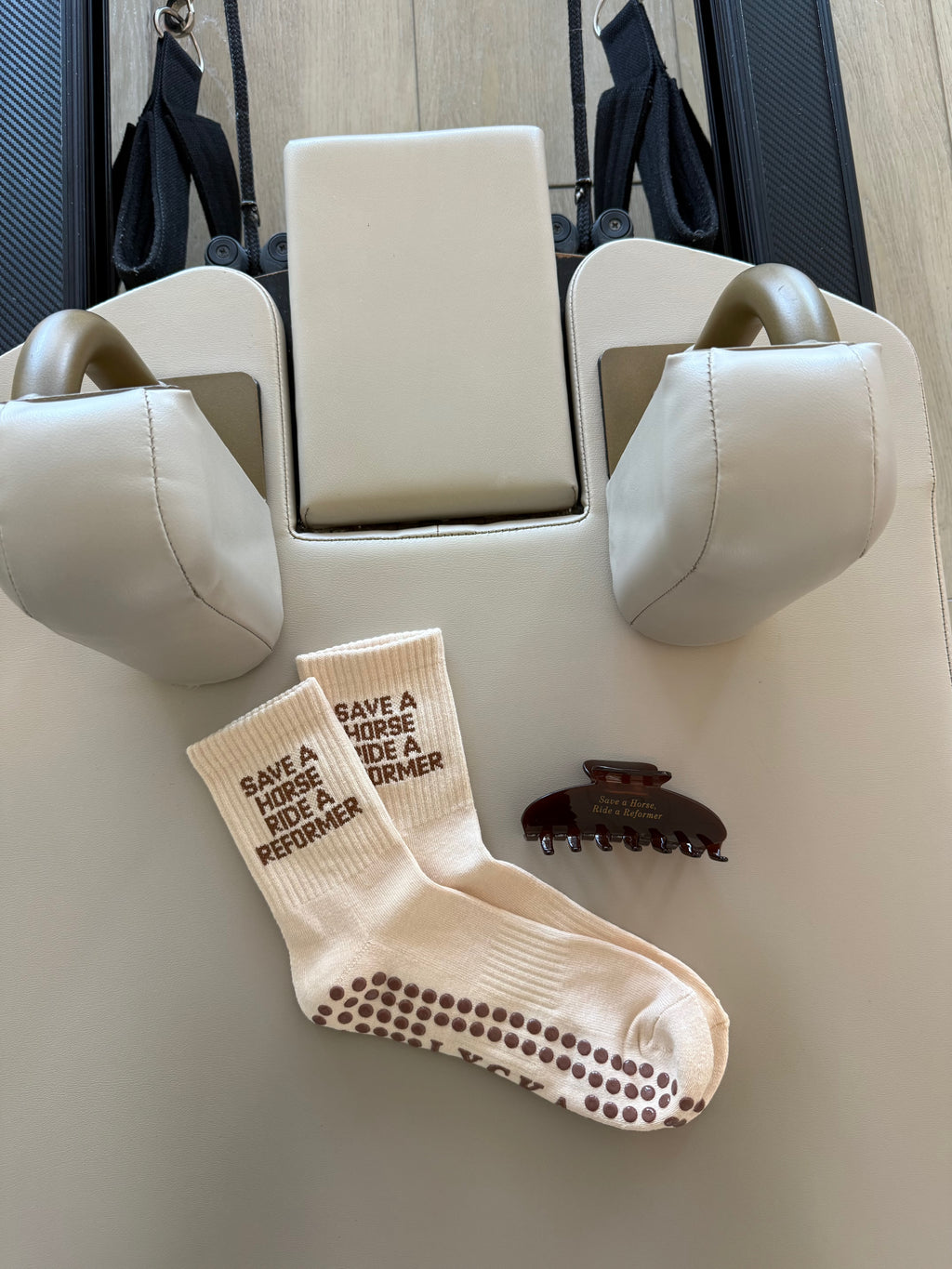 Beige Pilates socks saying “save a horse ride a reformer” and a brown hair clip that says “save a horse, ride a reformer” in gold font on a Pilates reformer 