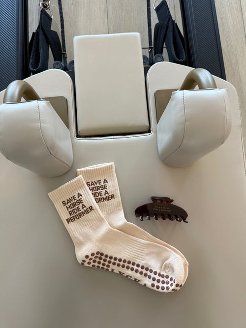 Beige Pilates socks saying “save a horse ride a reformer” and a brown hair clip that says “save a horse, ride a reformer” in gold font on a Pilates reformer 