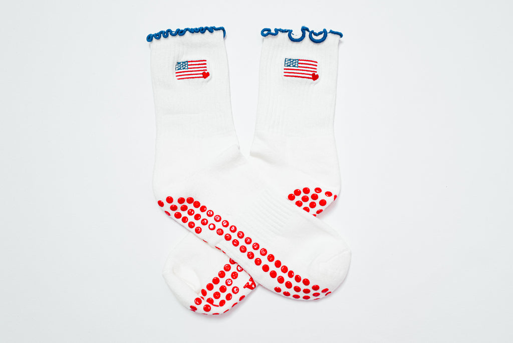 A pair of white grip socks with blue ruffle trim, a small American flag embroidered near the ankle, and red non-slip grip dots covering the soles. The socks are displayed crossed over each other on a white background.