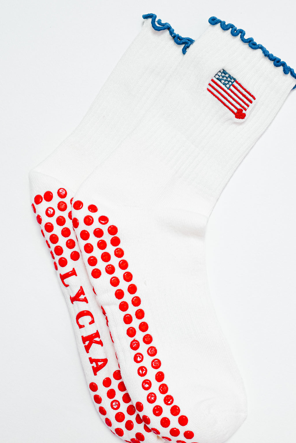 A pair of white grip socks with blue ruffle trim, a small American flag embroidered near the ankle, and red non-slip grip dots covering the soles. The socks are displayed layered over each other on a white background.