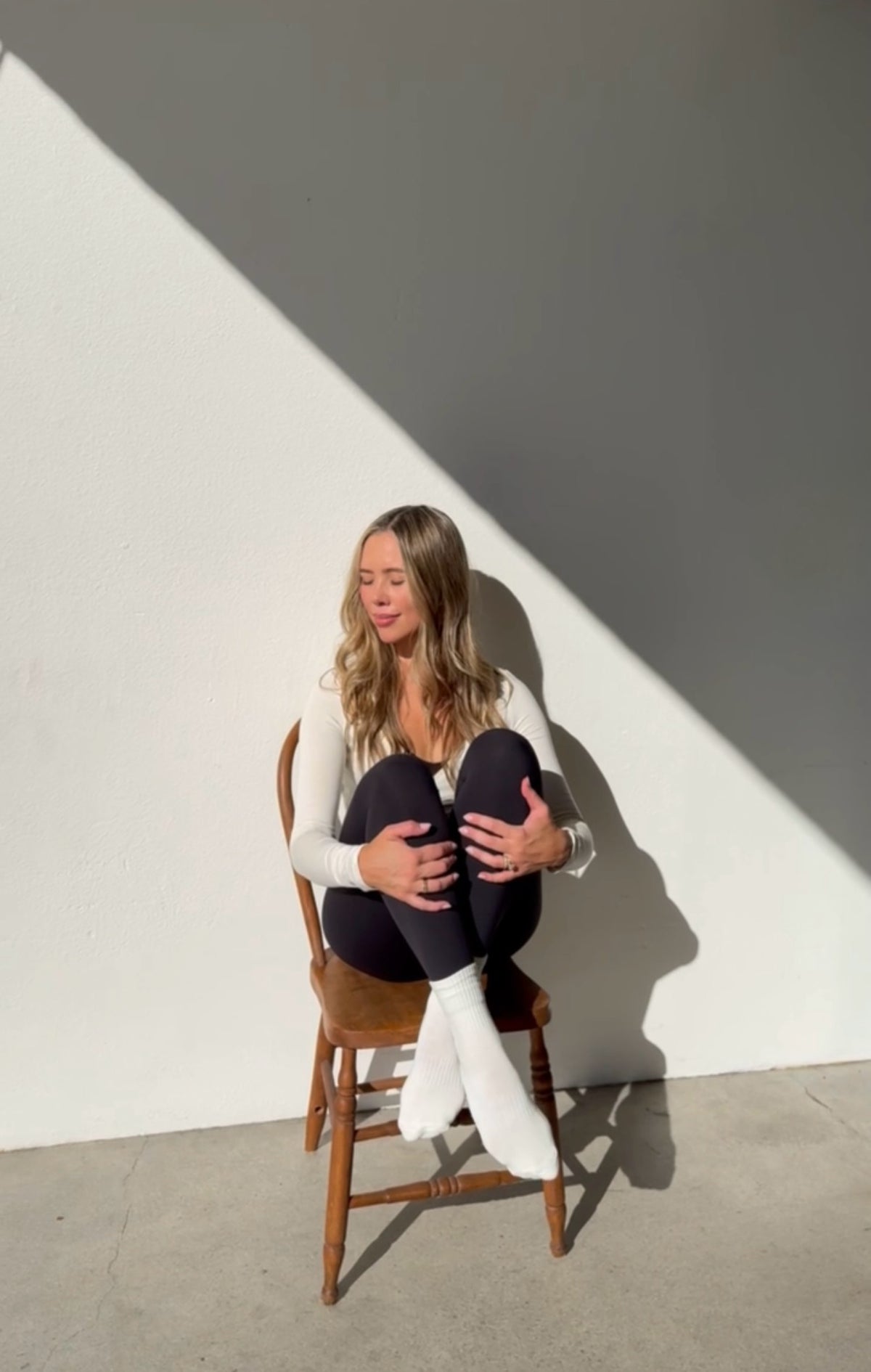 A woman sits on a wooden chair against a white wall with strong diagonal shadows, wearing a white long-sleeve top, black leggings, and white socks.