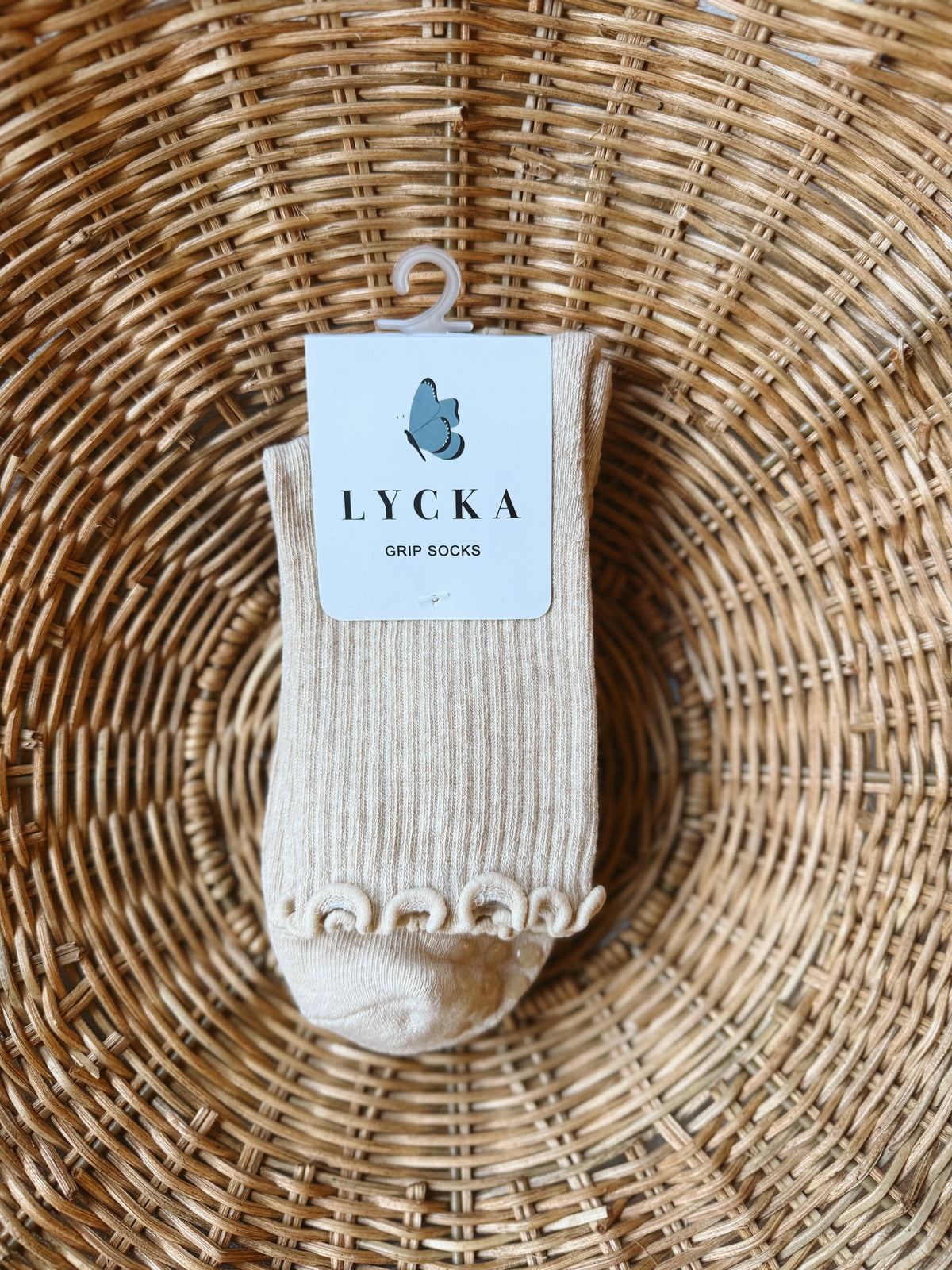 A single pair of LYCKA grip socks in a light beige/cream color, resting on a woven wicker tray. The socks feature a scalloped edge near the cuff and a white paper tag with the LYCKA brand logo.