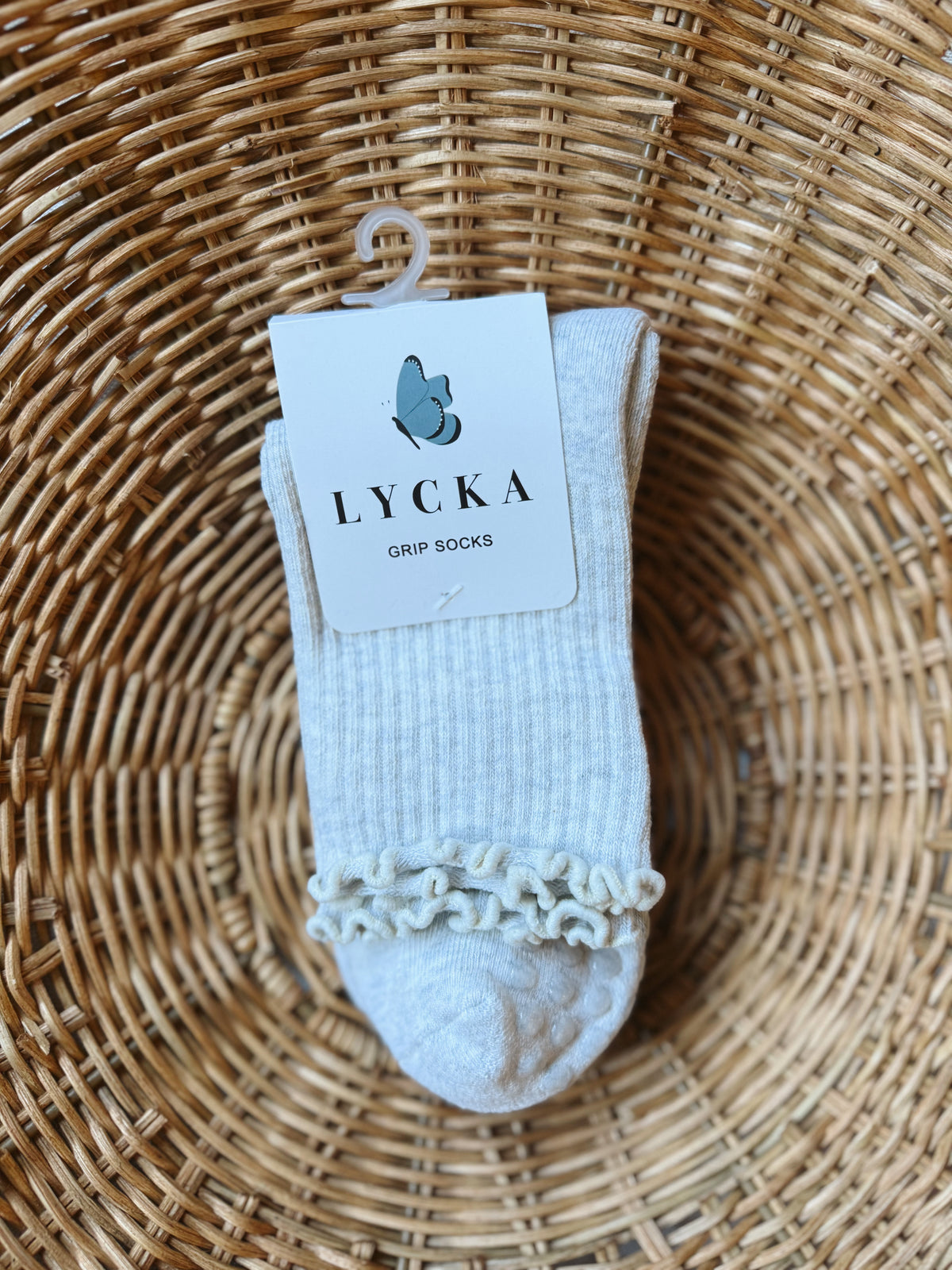 A pair of light gray ribbed grip socks with a decorative ruffled trim at the cuff, hanging from a white plastic hanger with a paper tag that reads 'LYCKA GRIP SOCKS'. The socks are displayed inside a round woven wicker basket.