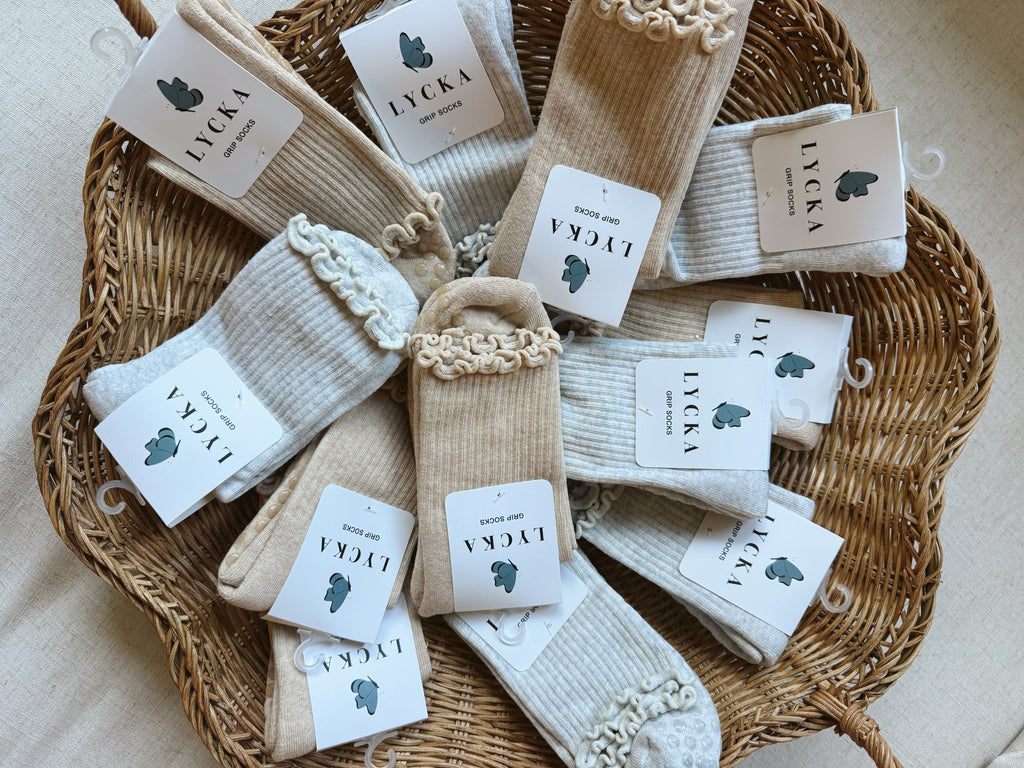 A styled flat-lay of multiple pairs of socks in neutral tones arranged in a round woven wicker basket. Each pair has a small paper tag with the brand name 'LYCKA' and a small dark blue butterfly logo.