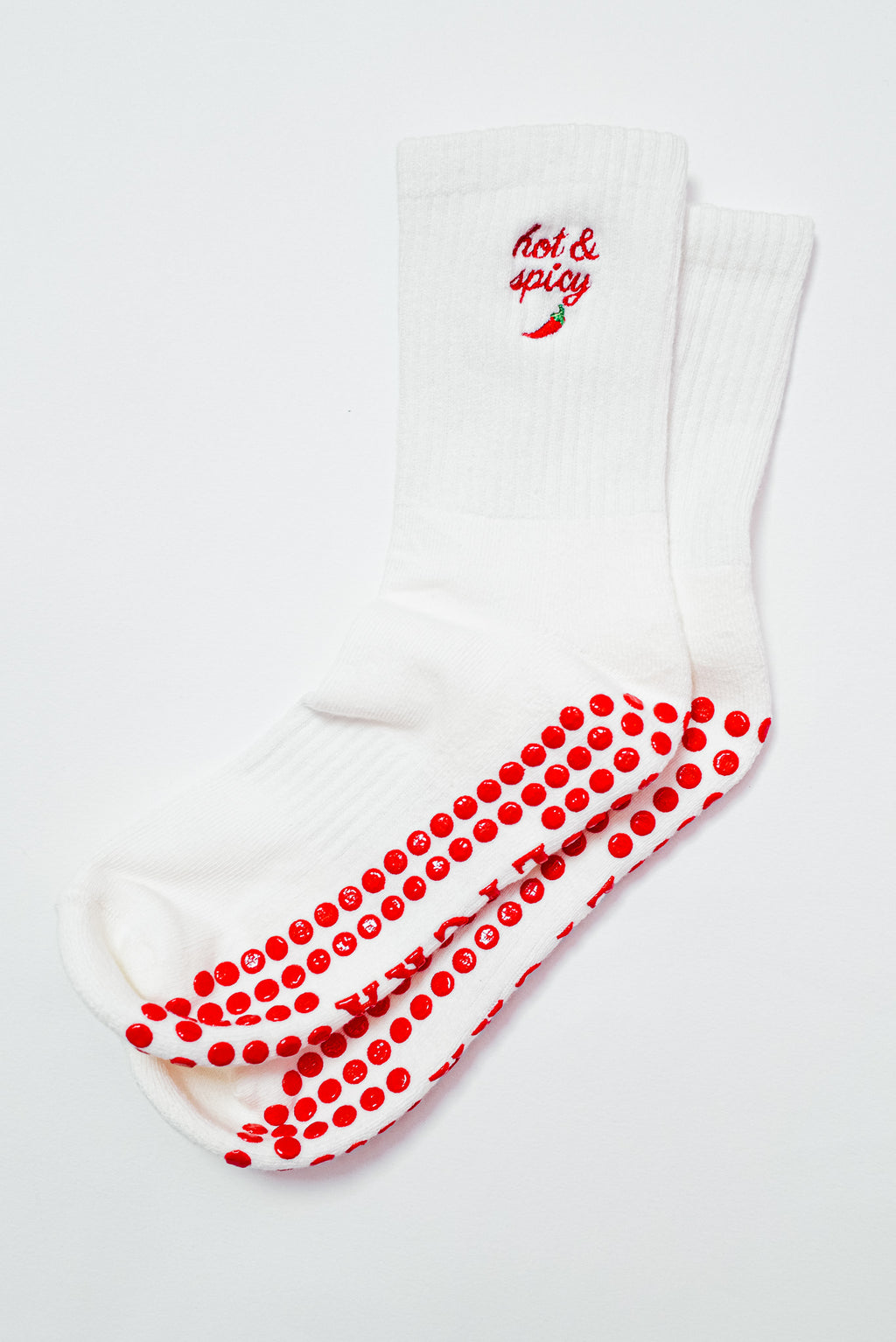 A pair of white grip socks displayed on a white background. The socks feature red grip dots on the bottom and a red embroidered "Hot & Spicy" design with a small chili pepper near the ankle.