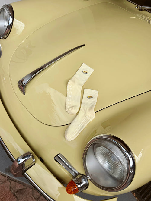 A pair of light yellow-colored textured socks with small butter embroidered details, laid flat on the glossy cream-colored hood of a vintage car.