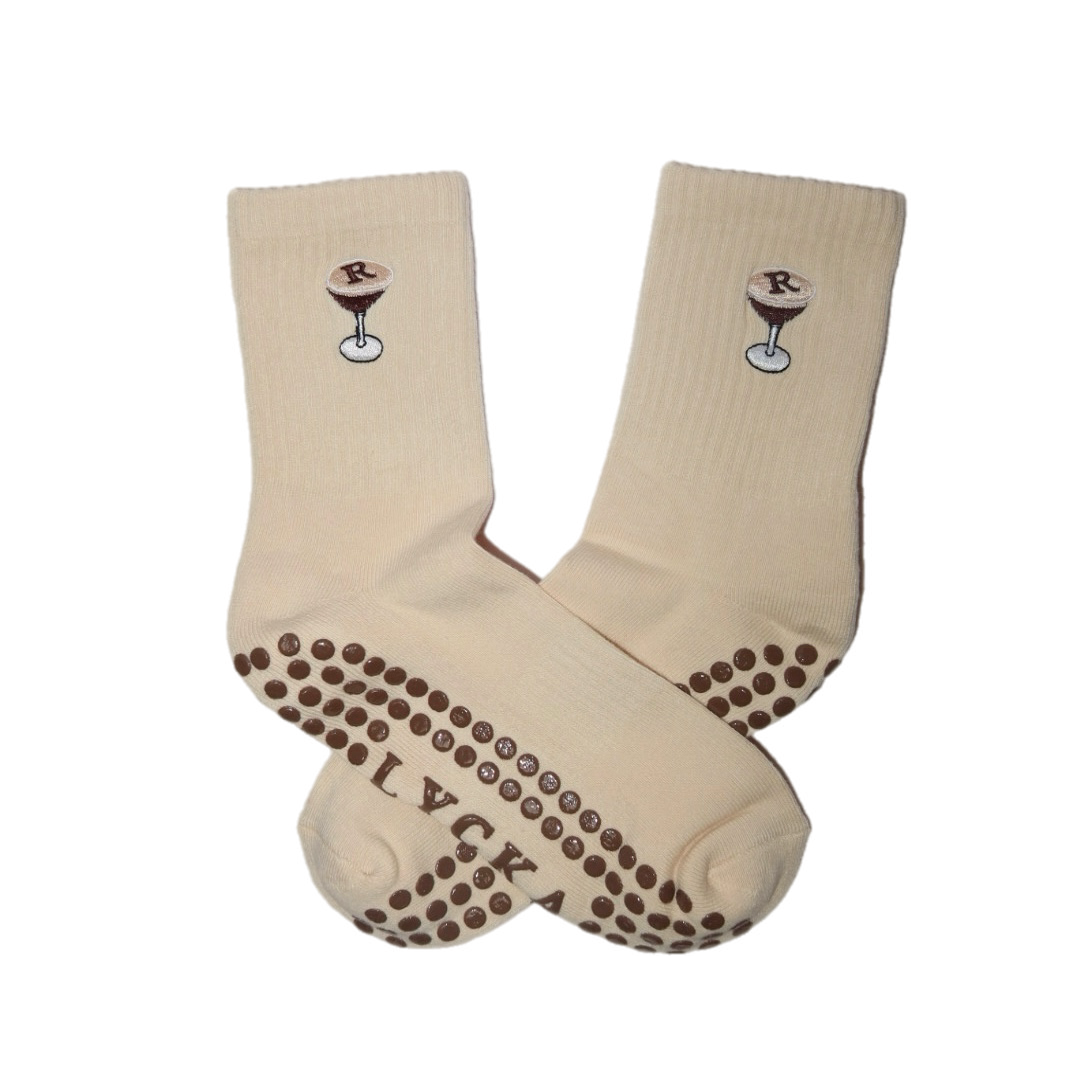 A pair of cream-colored grip socks with brown silicone dots on the sole and heel, featuring a small cocktail graphic with an 'R' on the side.