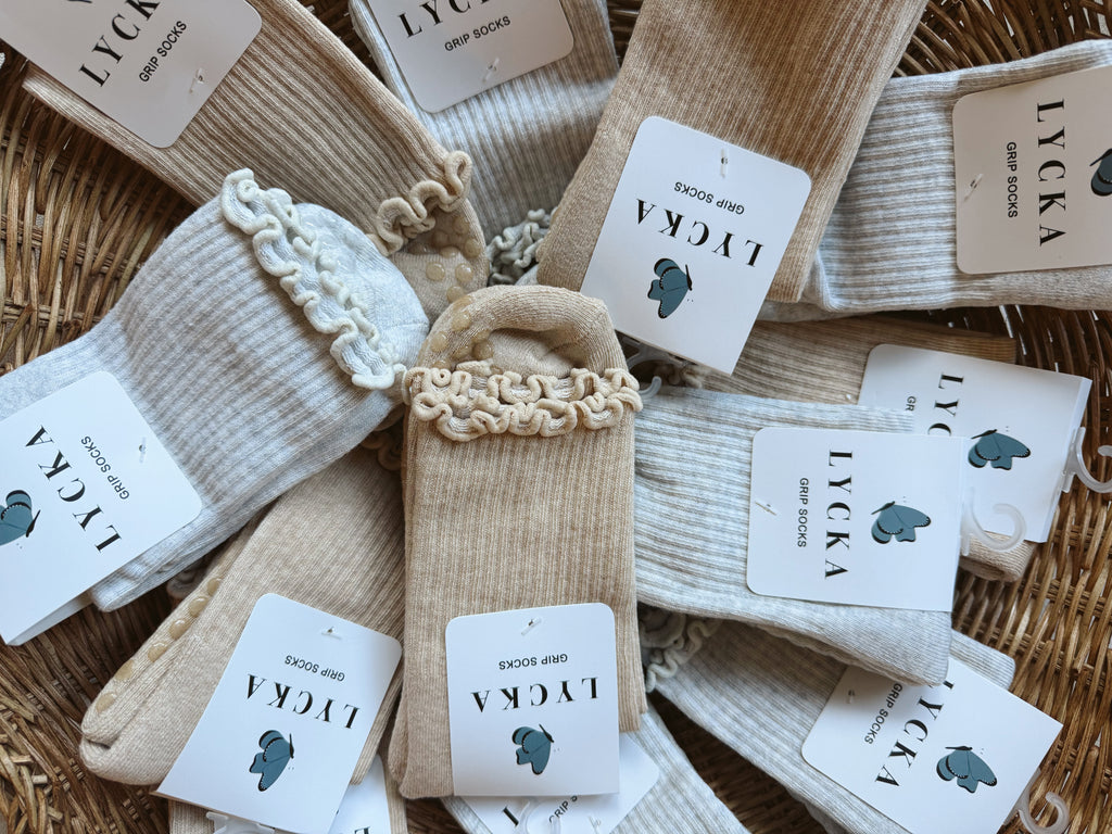 A styled flat-lay of multiple pairs of LYCKA grip socks in cream, beige, and light gray, arranged in a wicker basket. Each pair has a white paper tag with the brand name 'LYCKA' and a small blue butterfly logo.