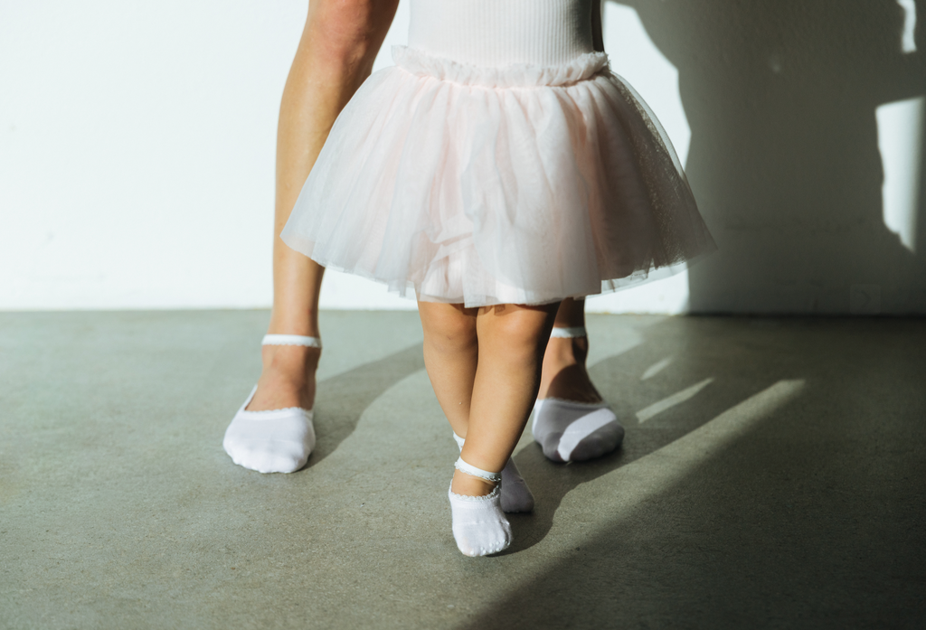 A close-up lifestyle shot of a child's legs in a baby pink tutu skirt and white ballet grip socks, standing on a concrete floor with a dancer's legs behind them in soft sunlight and shadows.