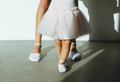 A close-up lifestyle shot of a child's legs in a baby pink tutu skirt and white ballet grip socks, standing on a concrete floor with a dancer's legs behind them in soft sunlight and shadows.